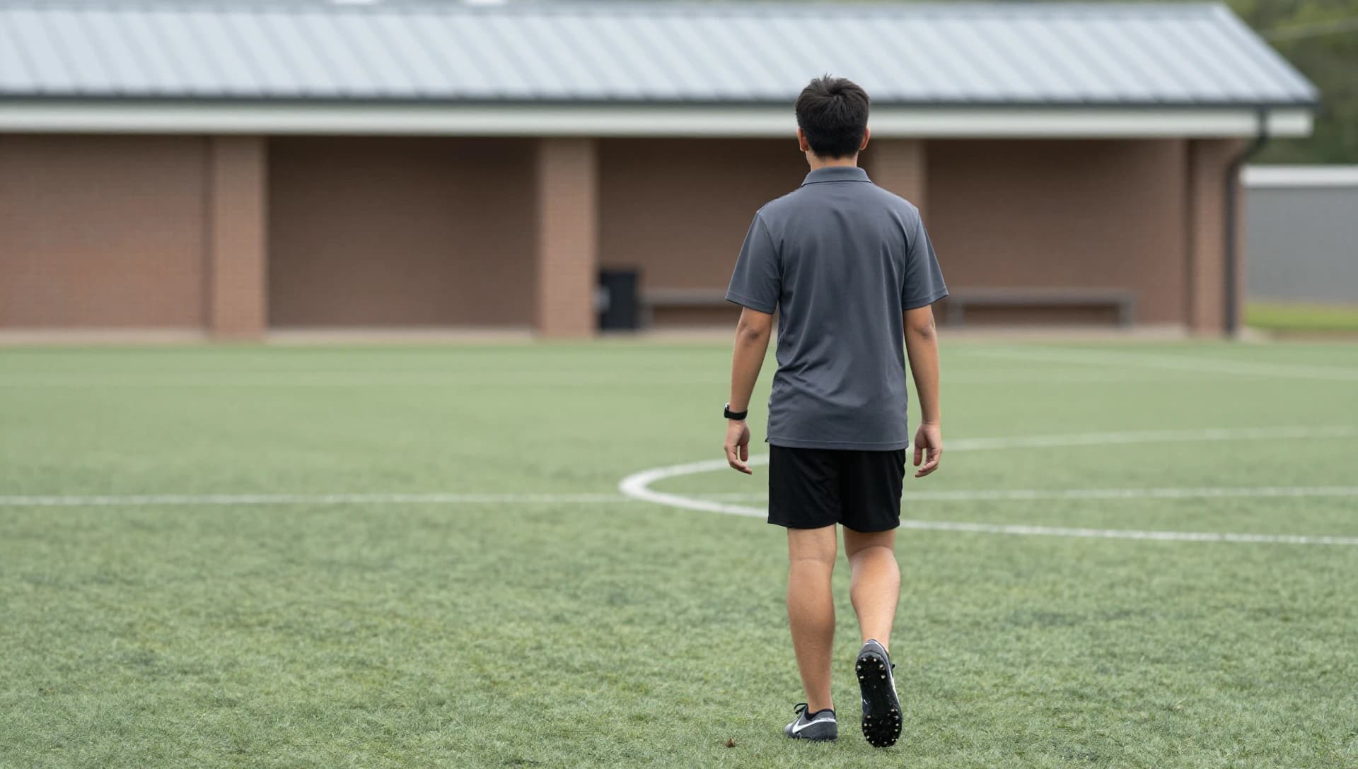 A youth soccer coach walking away across a green training field toward a distant clubhouse on a hazy morning, seen from behind.