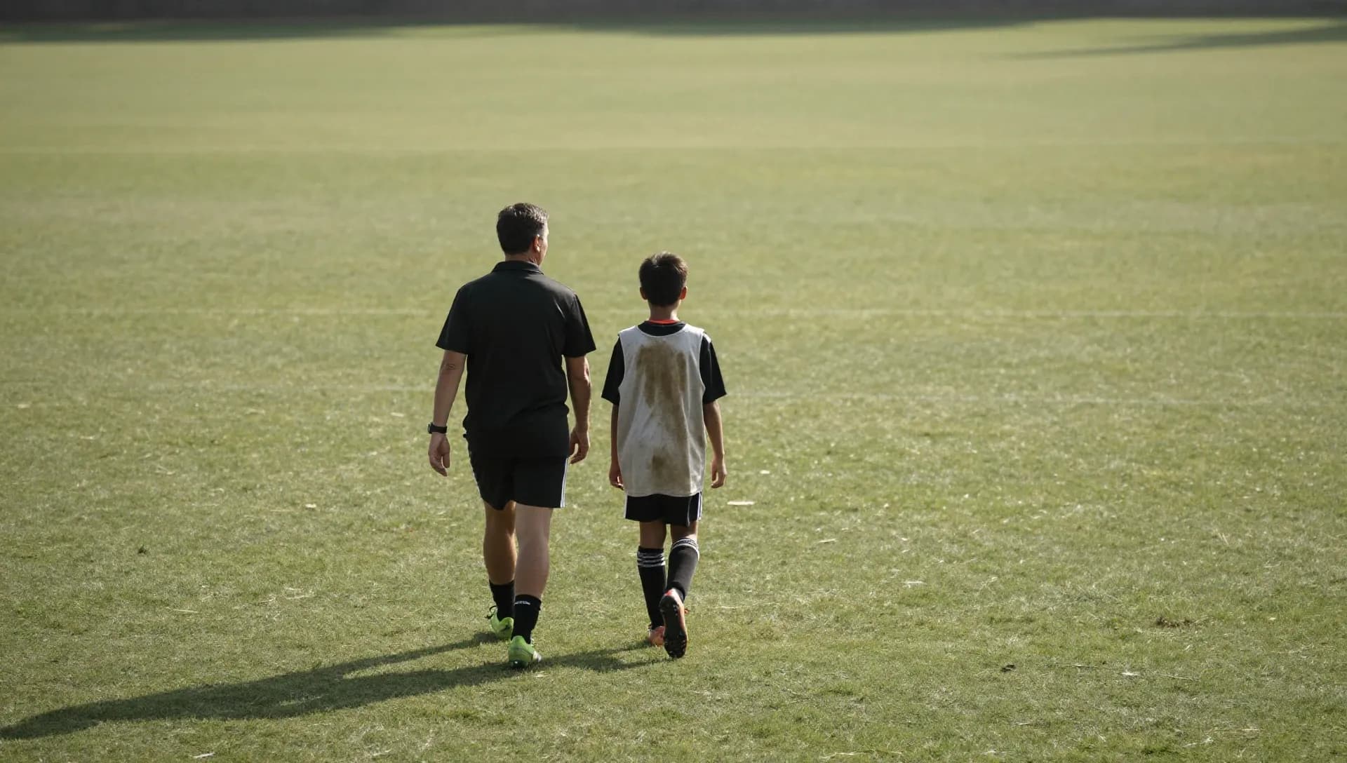 A youth soccer coach and a young player in a training bib walking together across a sunlit green pitch, seen from behind.